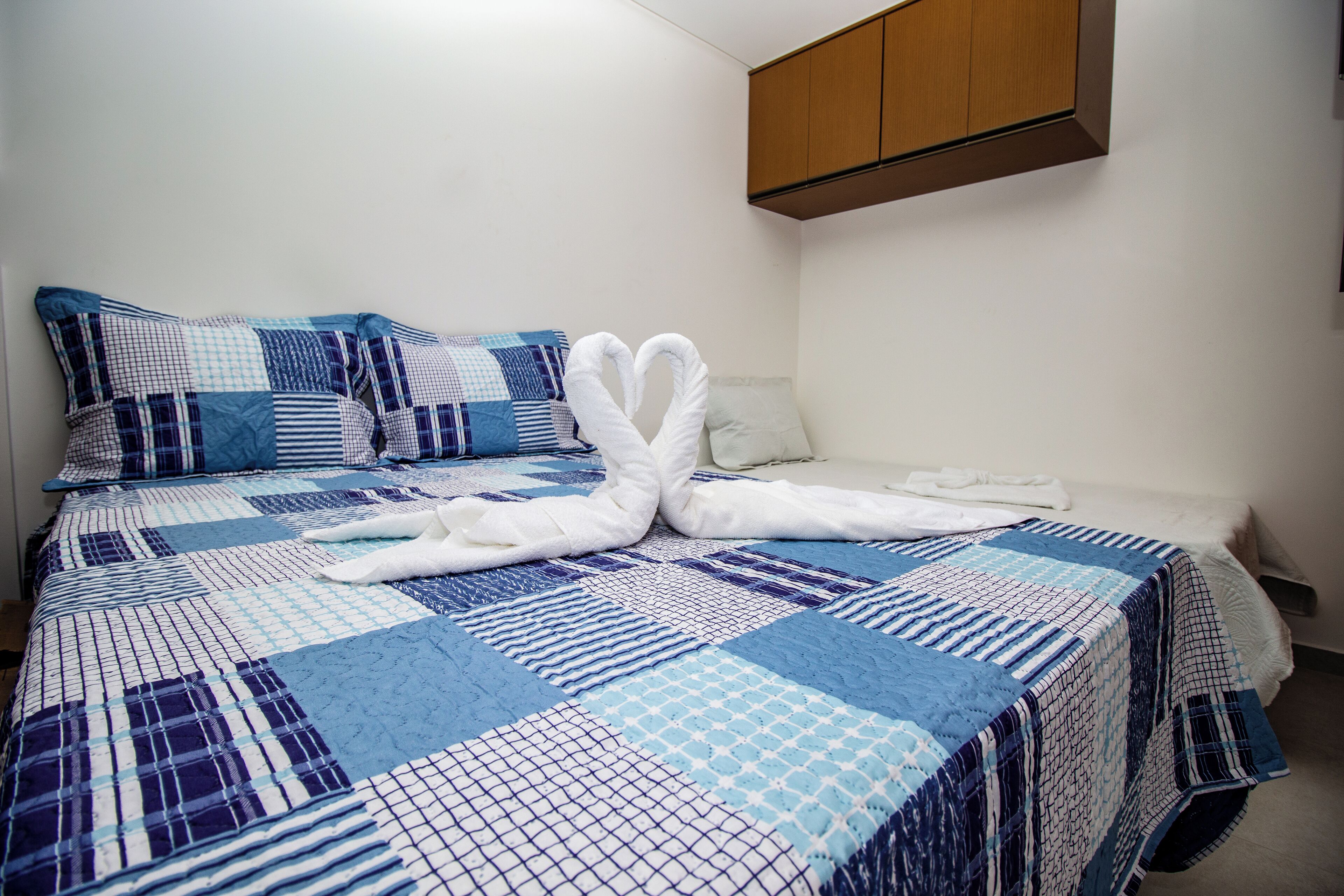 A neatly made bed with a blue and white quilt, two white towels arranged in a heart shape, and a wooden cabinet above the bed.