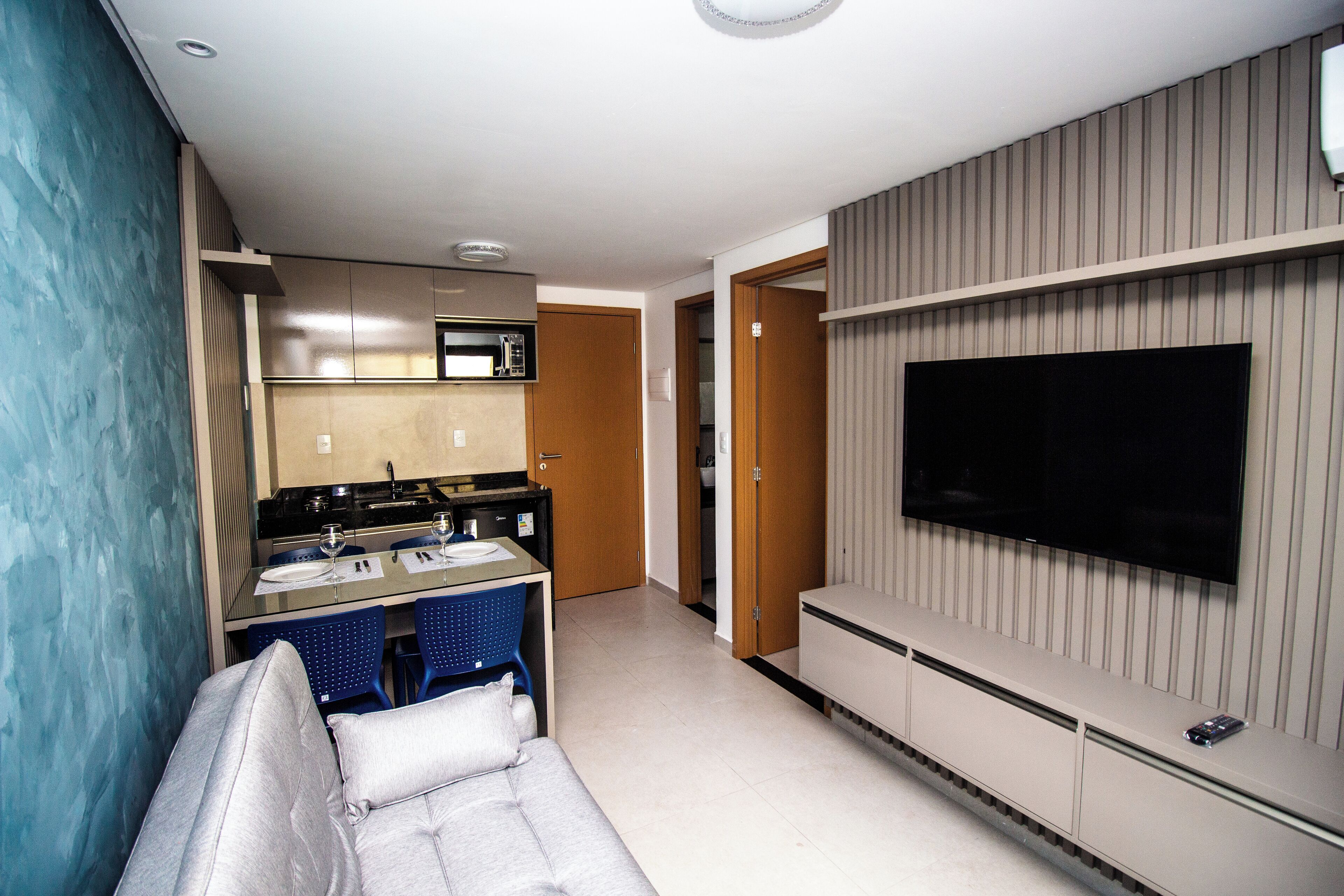 A compact living space with a kitchenette, a sofa, a TV mounted on the wall, and a door leading to another room.