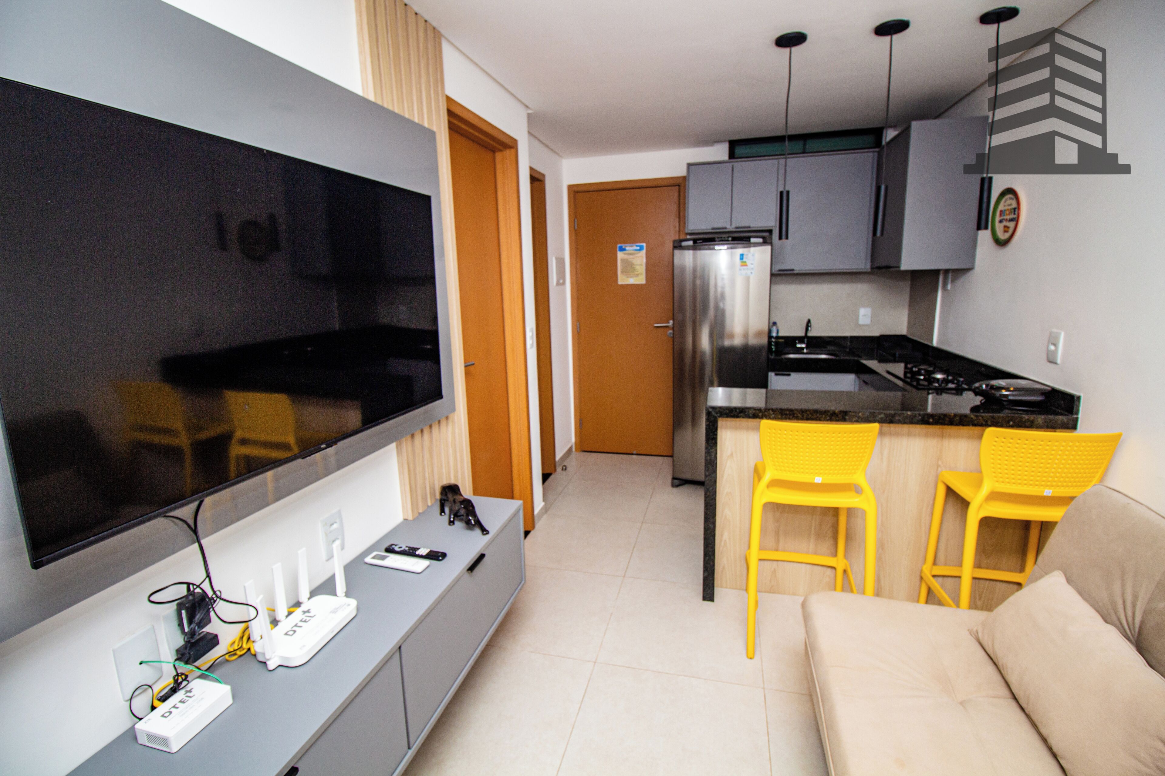 Una cucina moderna con frigorifero, piano cottura e sgabelli da bar. C'è una TV a schermo piatto montata a parete e un divano nella zona soggiorno.