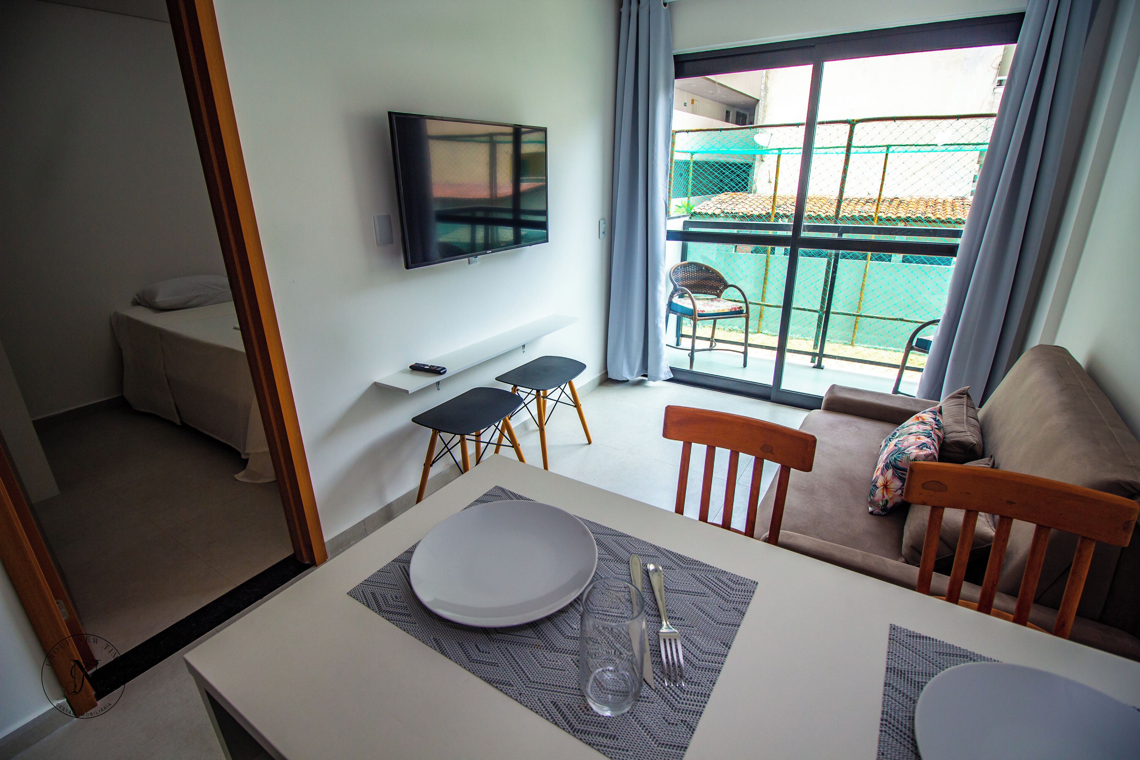 A dining area with a table set for two, a sofa, a TV mounted on the wall, and a view of the outside balcony.