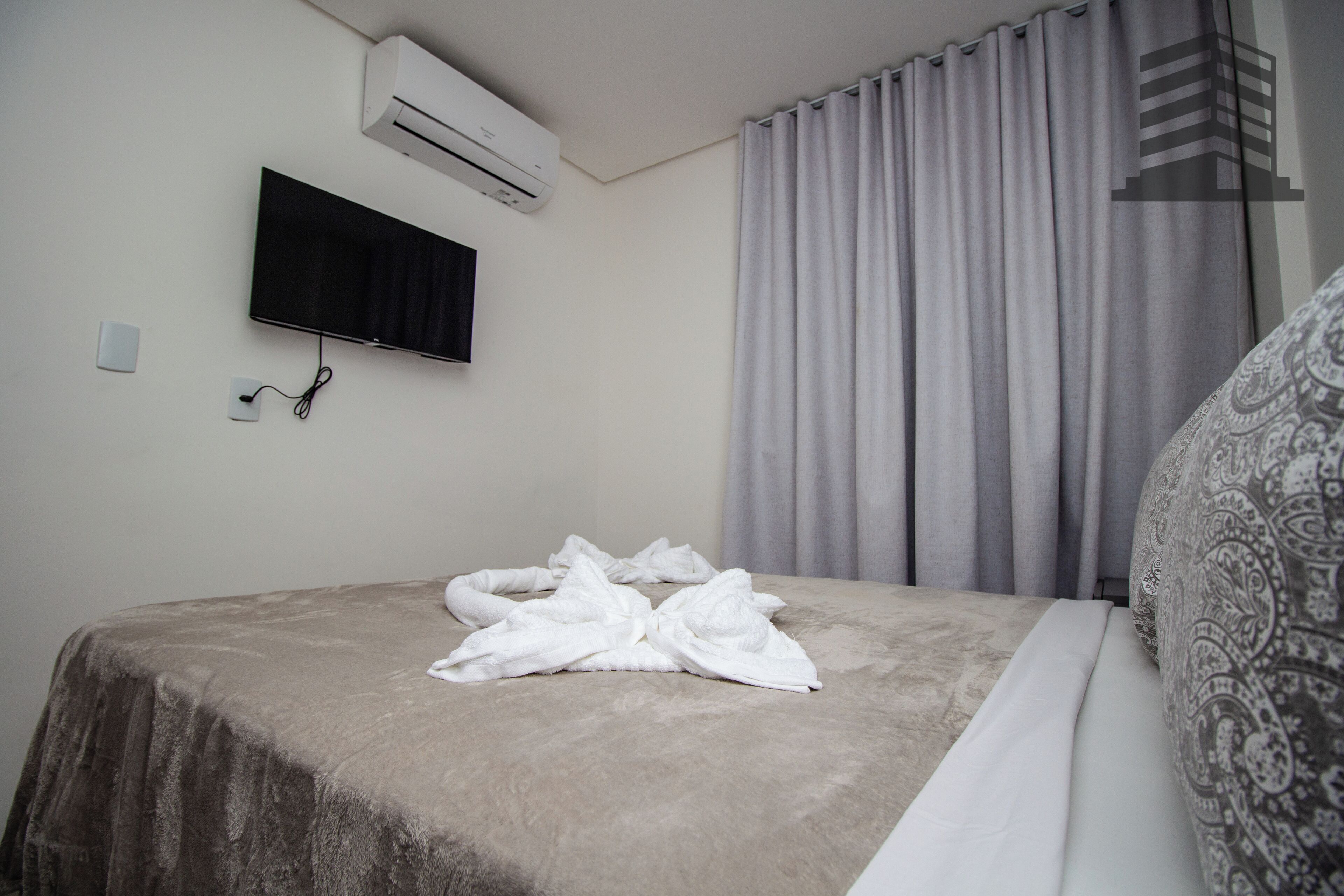 Una cama bien hecha con una manta gris, un televisor de pantalla plana montado en la pared y una ventana con cortinas grises.