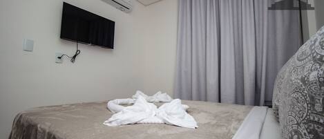 Una cama bien hecha con una manta gris, un televisor de pantalla plana montado en la pared y una ventana con cortinas grises.