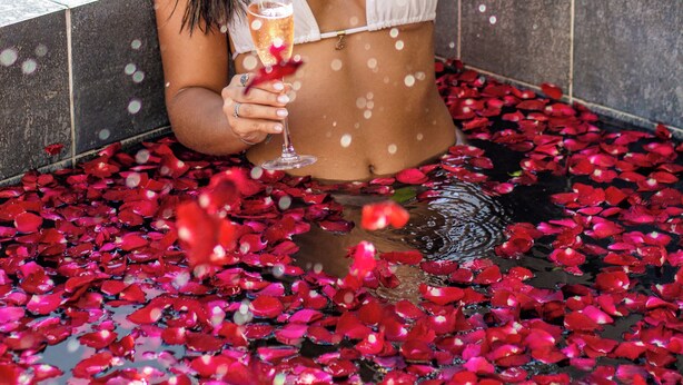 A person in a white bikini sitting in a pool filled with rose petals, holding a glass of champagne. - Bela Vista Beach By New Time (Ipojuca)