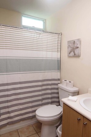 A bathroom with a striped shower curtain, a toilet, a sink with a towel, and a starfish decoration on the wall. - #18 First floor 3br, 2ba Apartment @ Jobos Beach (Isabela)