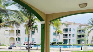 A covered outdoor seating area with a pool view, wooden furniture, and a building with balconies in the background. - #18 First floor 3br, 2ba Apartment @ Jobos Beach (Isabela)
