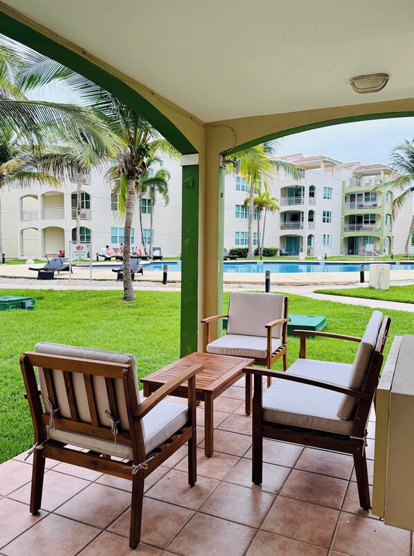 A covered outdoor seating area with a pool view, wooden furniture, and a building with balconies in the background. - #18 First floor 3br, 2ba Apartment @ Jobos Beach (Isabela)