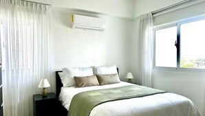 Une chambre à coucher comprenant un lit, deux tables de chevet, un climatiseur et une fenêtre avec des rideaux.