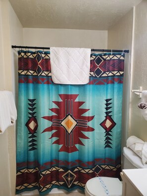 A bathroom with a shower curtain featuring a geometric pattern, a white towel hanging on the curtain rod, and a toilet with a white seat. - Southwest Motel (Grants)