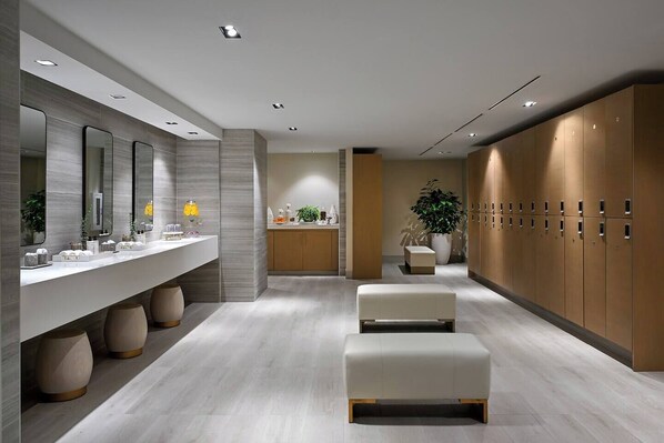 Un baño moderno con un mueble de tocador amplio, varios espejos, un banco, taquillas y una planta en maceta.