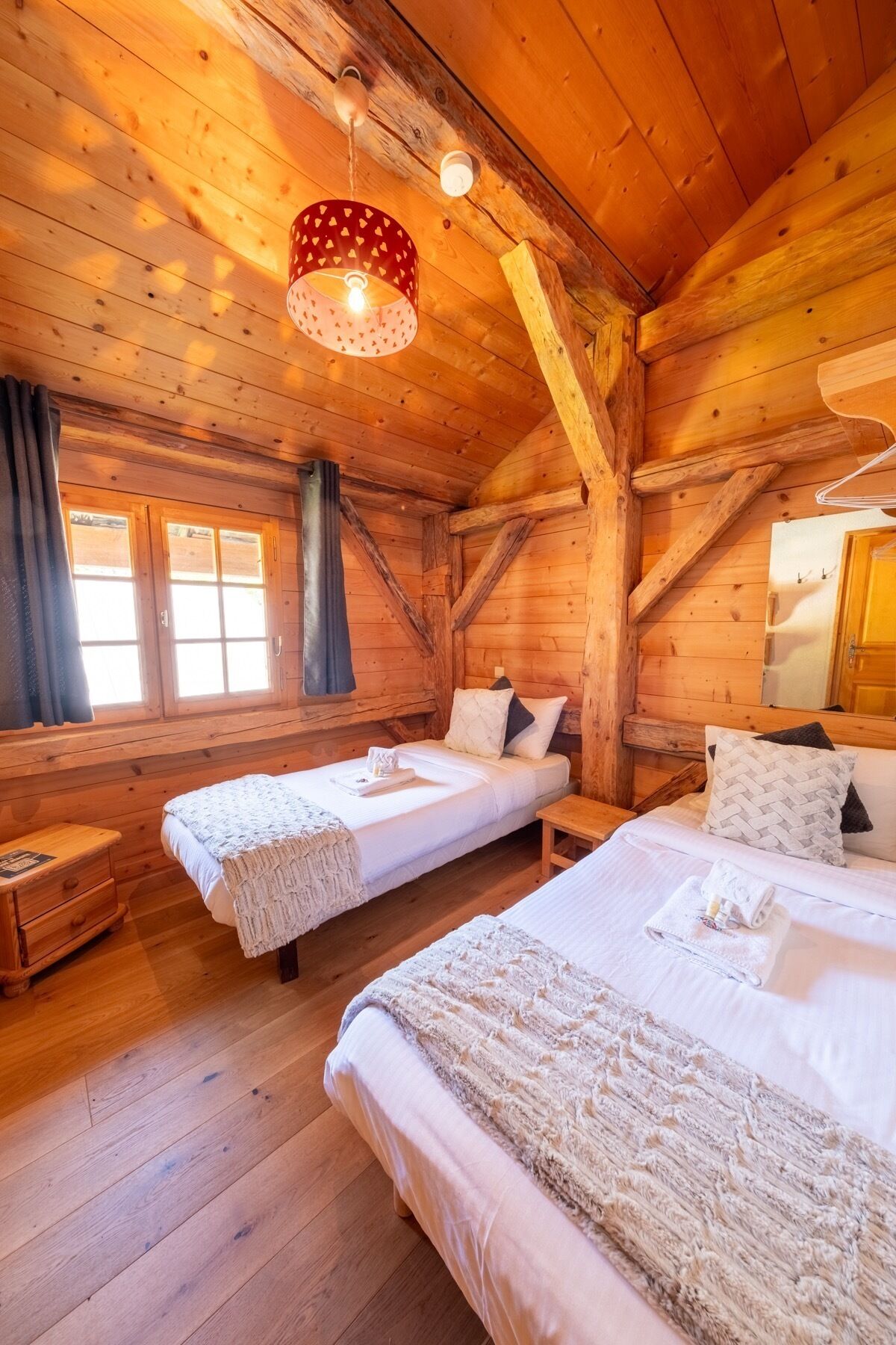 Une chambre confortable, aménagée dans le style d’un chalet, avec deux lits simples, des murs en bois, un plafond en pente et une fenêtre avec des rideaux.