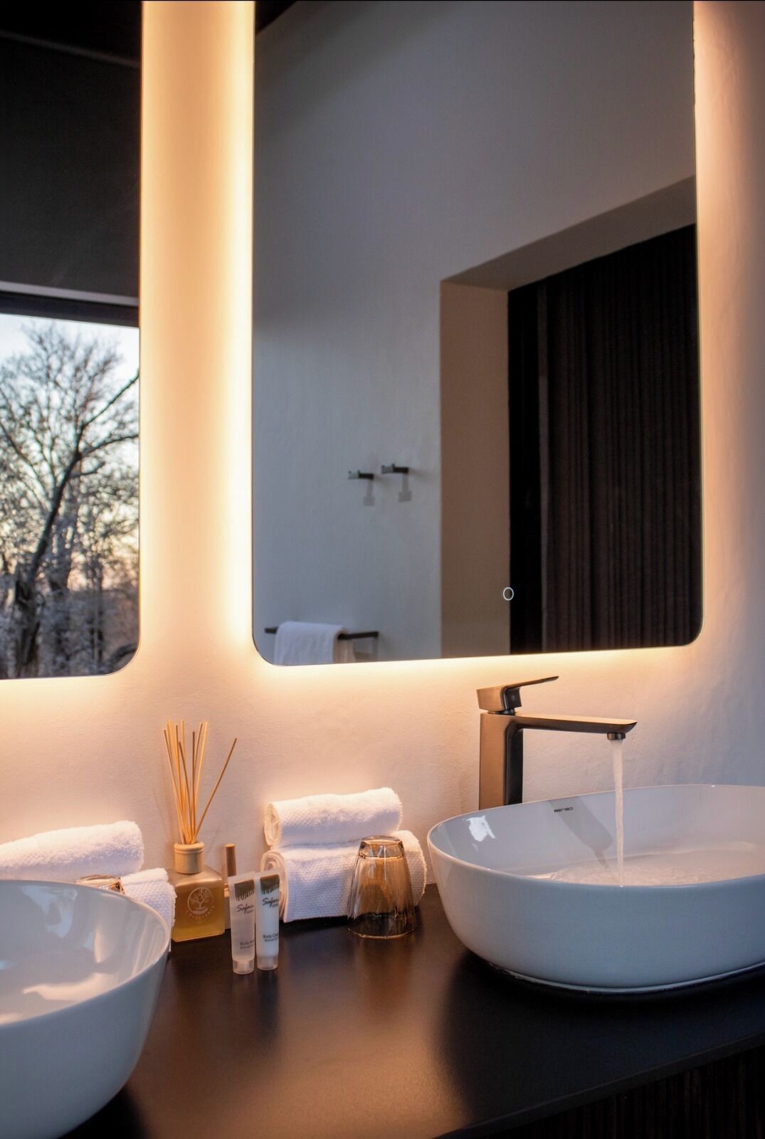 A modern bathroom with two large white sinks, a mirror, and a window with a view of trees.