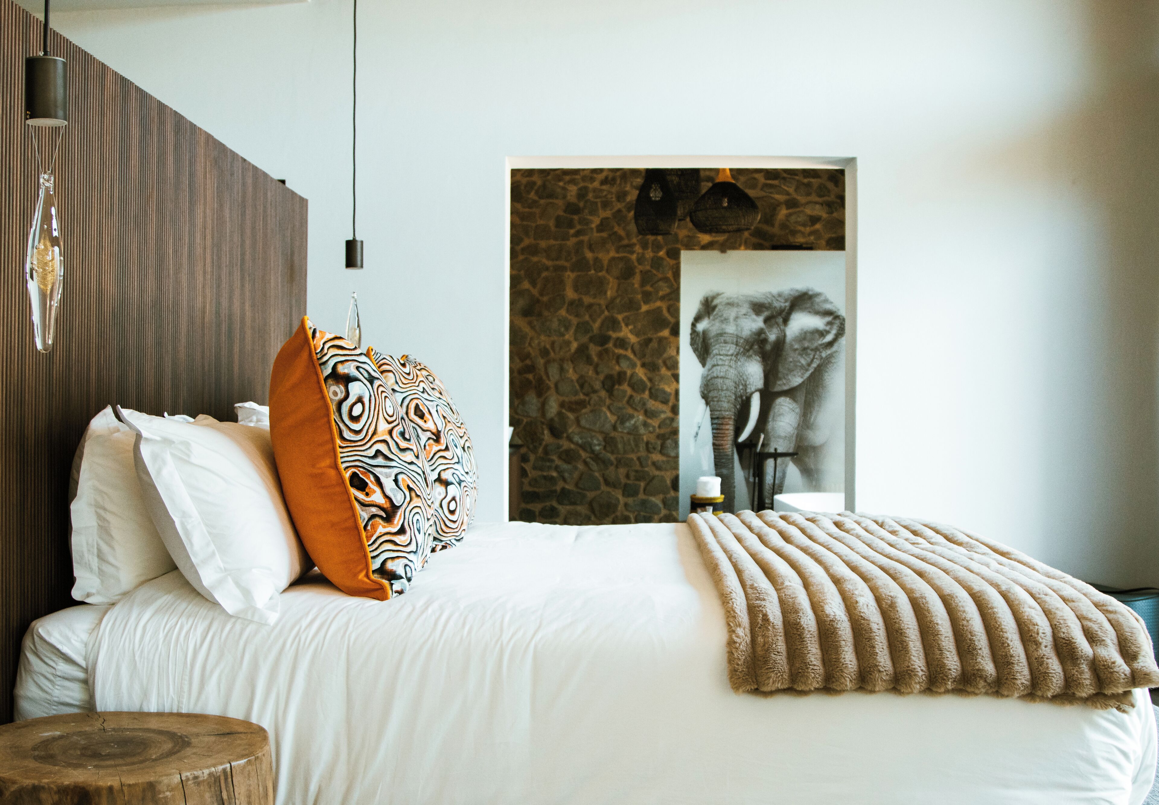 A bedroom with a bed, pillows, a blanket, a wooden stool, and a wall with a stone pattern and a framed picture.