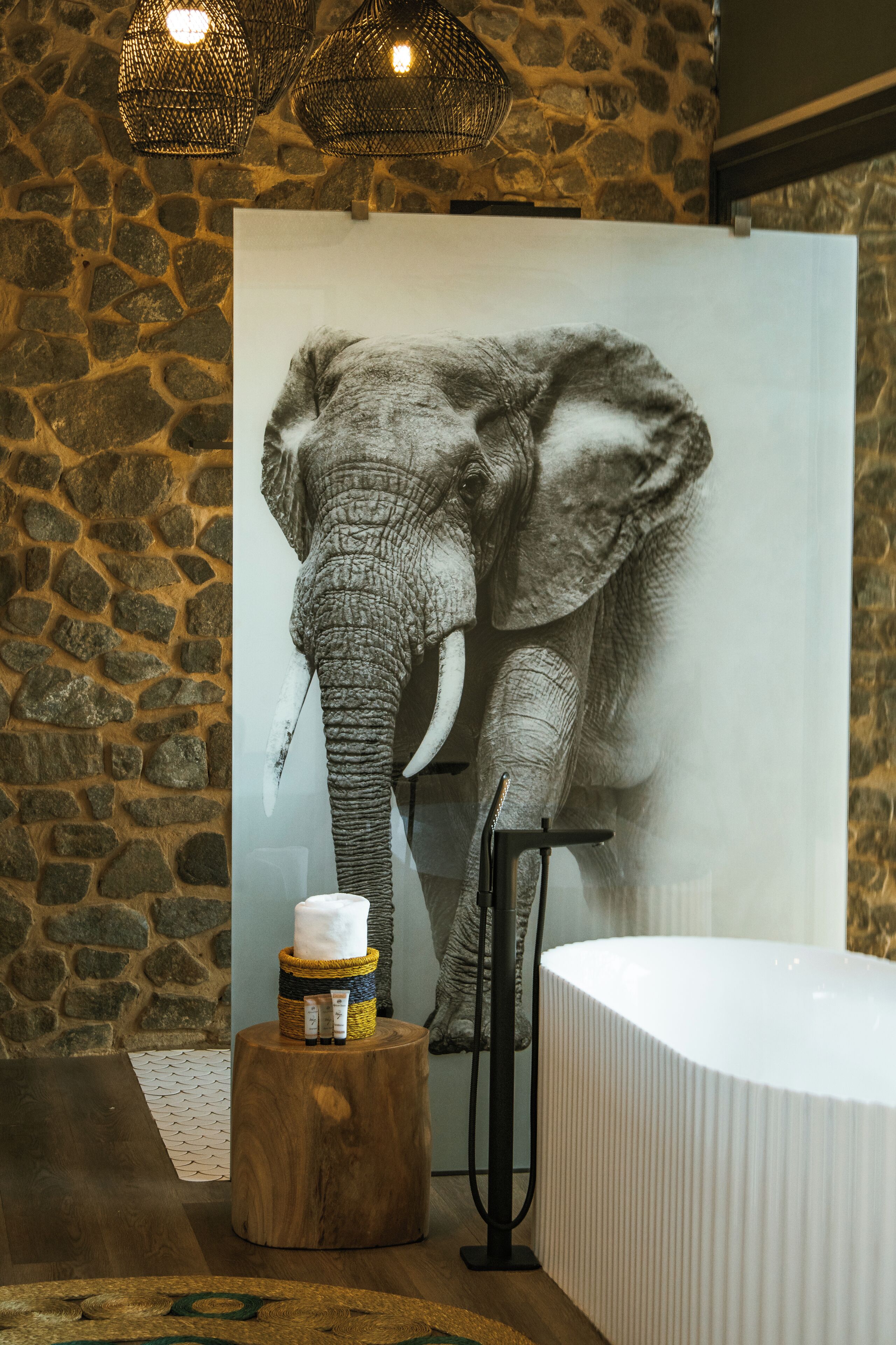 A bathroom with a stone wall, a large elephant mural, a wooden stool with toiletries, and a white bathtub.