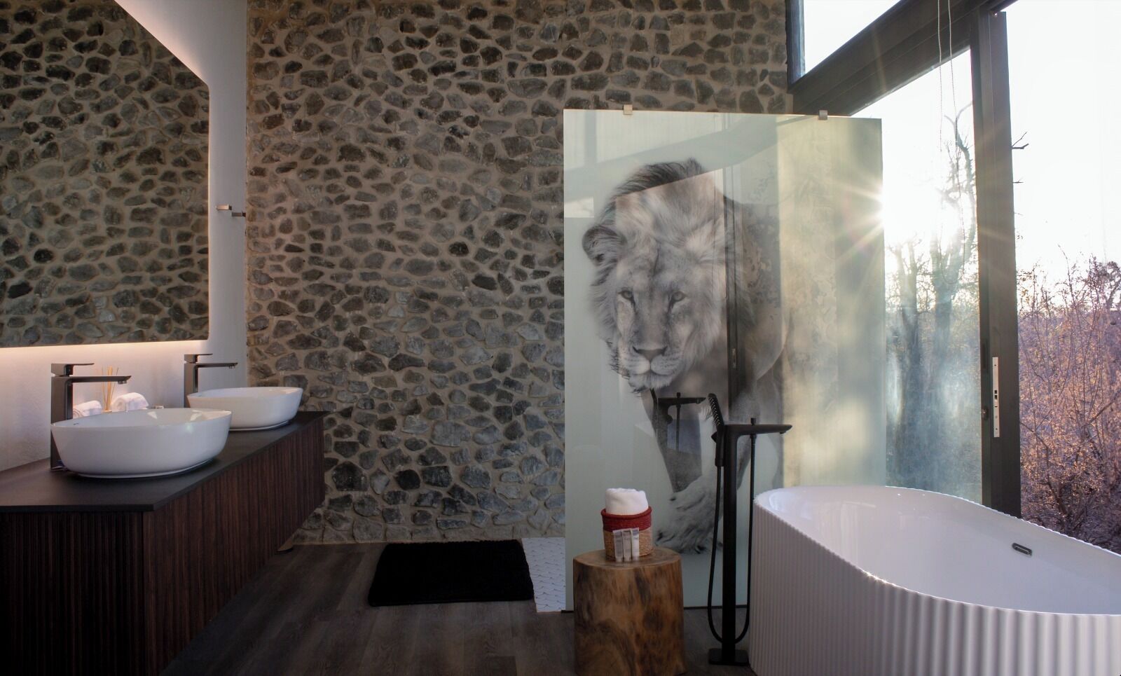 A modern bathroom with stone walls, two sinks, a large mirror, and a glass shower enclosure.