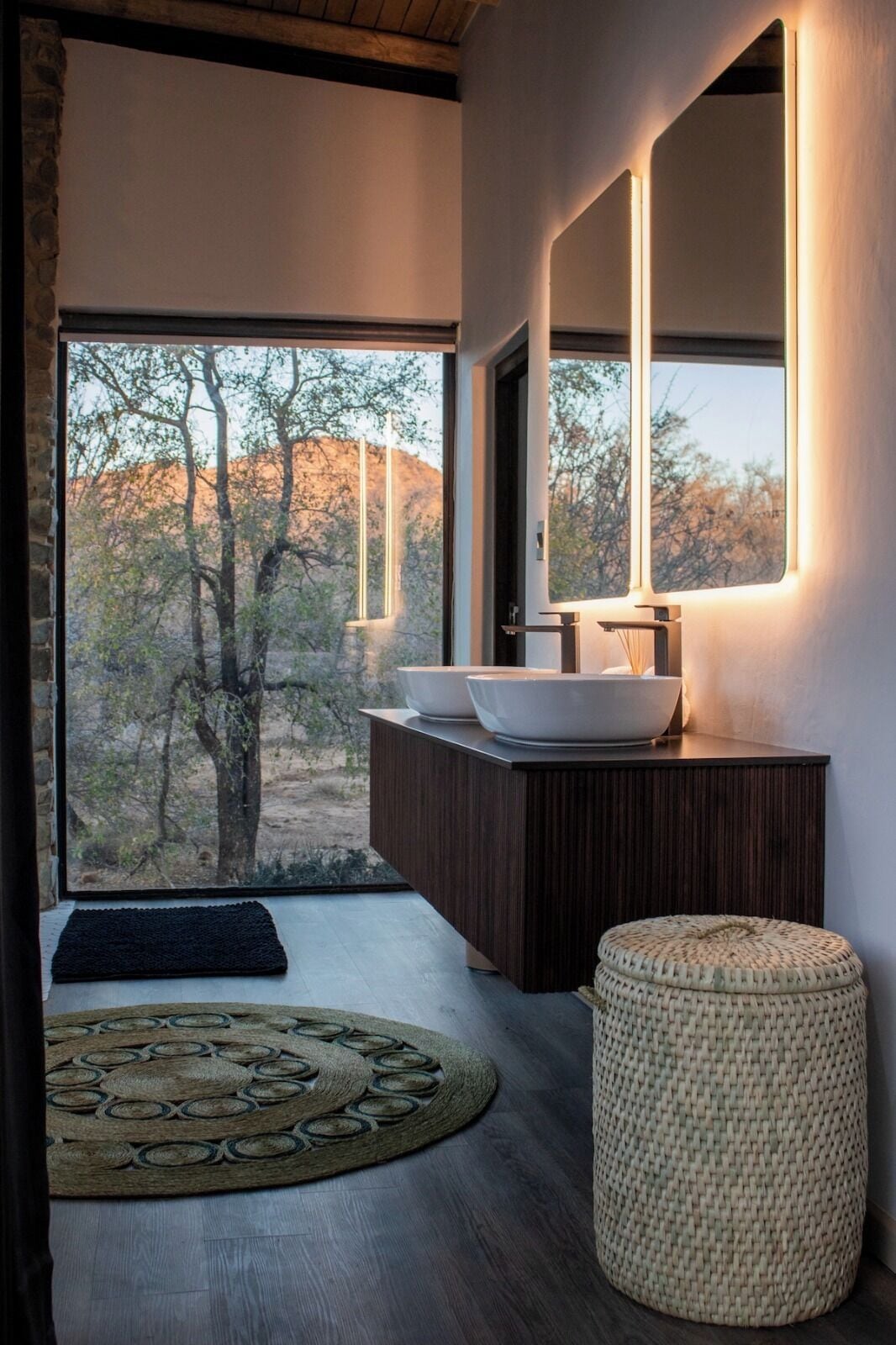 A modern bathroom with a large window, a wooden vanity, a round woven ottoman, and a decorative rug.