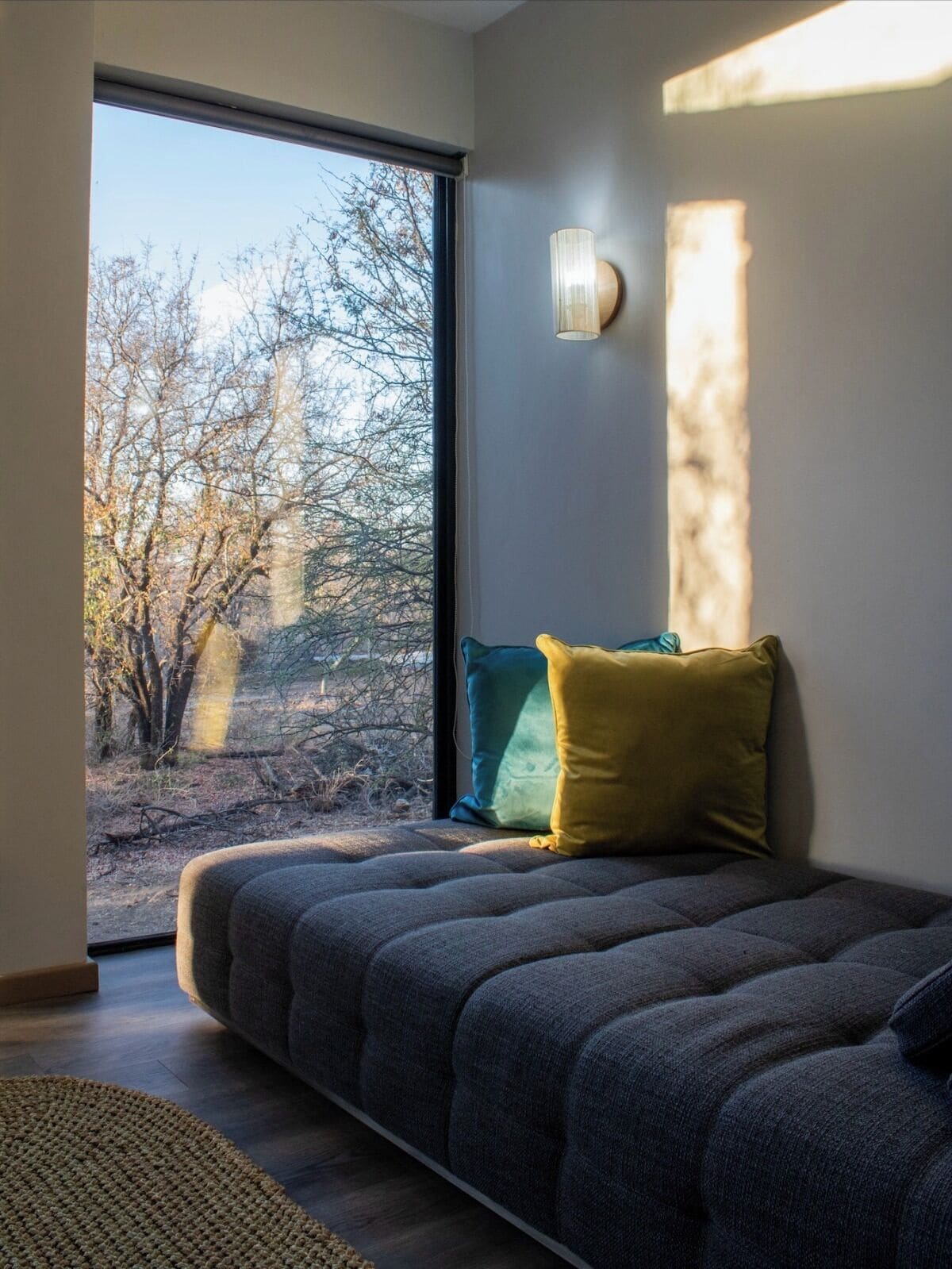 A modern bedroom with a large window, a bed with grey bedding, and a wall-mounted light fixture.