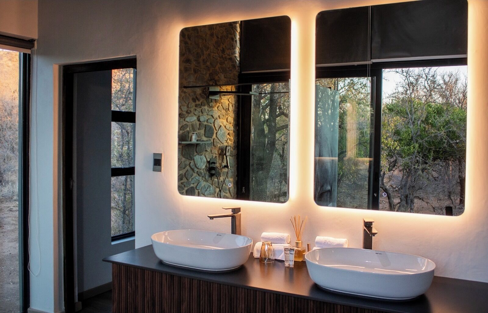 A modern bathroom with two large sinks, a stone accent wall, and a view of trees through the windows.