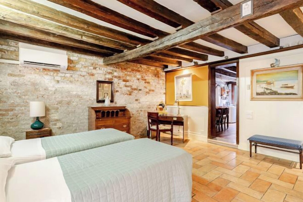 Una habitació amb llit, escriptori, cadira, banqueta i paret de maó.