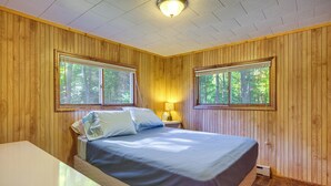 A wooden-paneled room with a bed, two windows, a nightstand, and a table. - 2 Blocks to Boat Ramp: Higgins Lake House w/ Grill (Roscommon)
