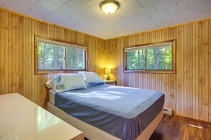 A wooden-paneled room with a bed, two windows, a nightstand, and a table.