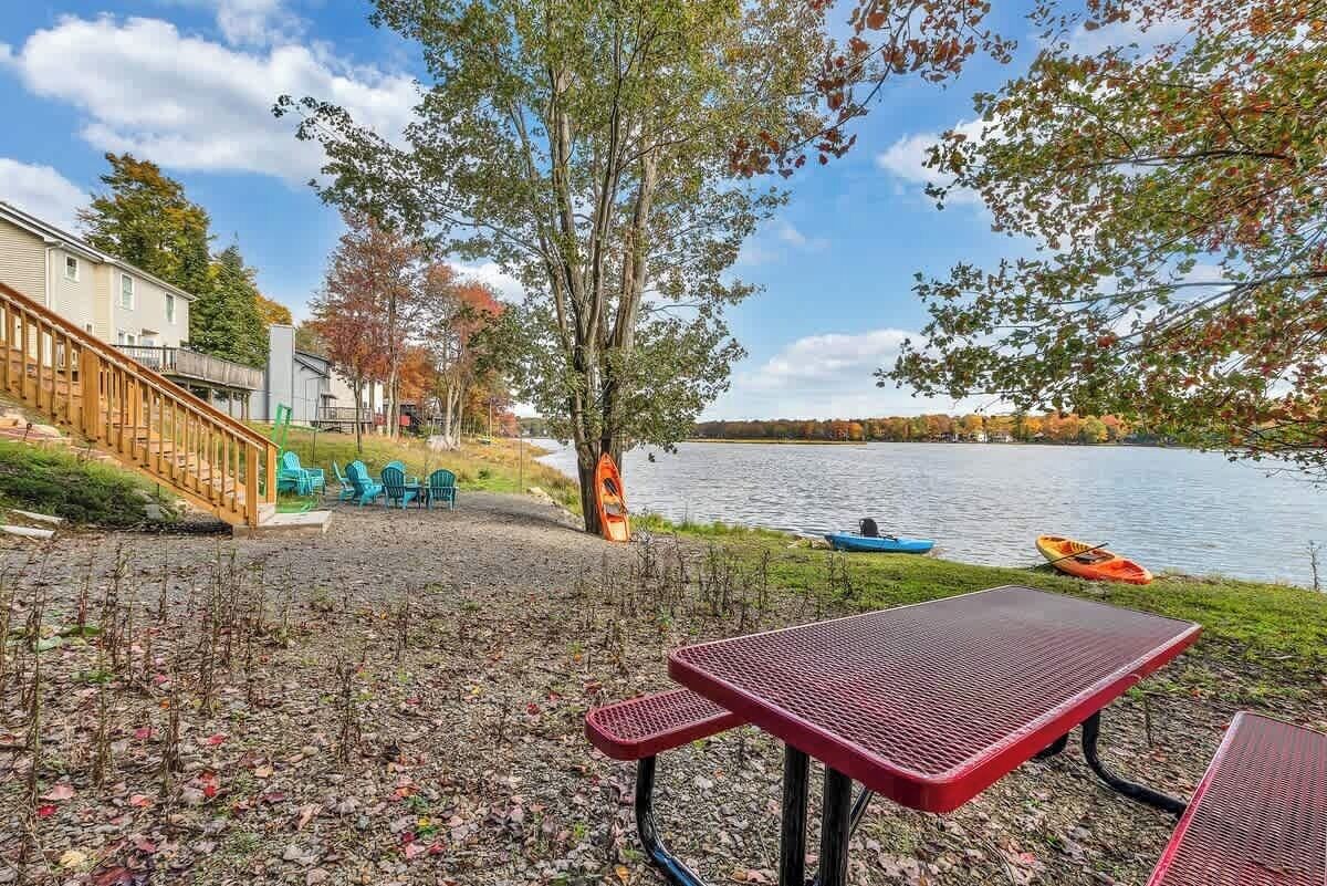 Un patio junto al lago con una mesa y sillas de metal rojo, una terraza de madera y una fila de sillas azules.