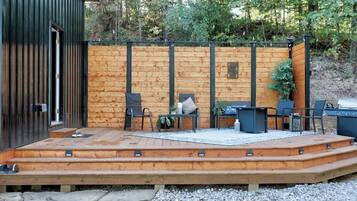 Una terraza de madera con mobiliario exterior, una parrilla y un sendero de piedra que conduce a un edificio.