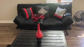 A black coffee table with a red vase in the center, a black sofa with patterned cushions, and a window with blinds in the background. - Charming 1-bedroom house rain Stone Mountain with WiFi access (Stone Mountain)