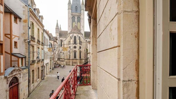 Une rue étroite bordée de bâtiments historiques, une église dotée d’un haut clocher et une rambarde rouge.