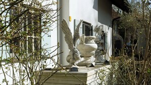 A statue of an angel with wings on a pedestal, surrounded by bare branches and a building in the background.