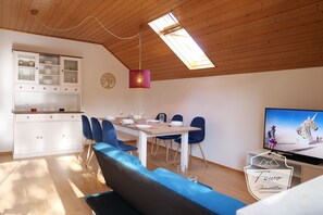 A dining area with a wooden ceiling, a dining table set with plates and glasses, blue chairs, and a television displaying a desert scene. - FeWo Schönwetter - Above (Weßling)