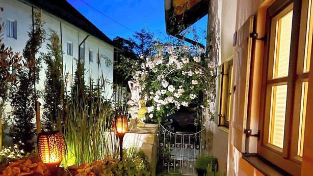 A narrow pathway flanked by white buildings, a decorative lamp, and a flowering bush.