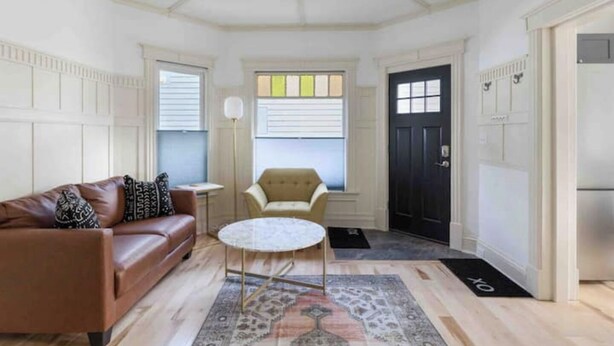 Una sala de estar con un sofá de cuero marrón, una mesa de centro redonda, un sillón color beige y una puerta negra.
