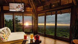 A room with a wooden floor, a white sofa, a table with drinks, and a TV displaying a couple. - Cabañas Cochapunko I Espectacular vista 360 (Salango)
