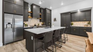 A modern kitchen with dark cabinetry, a stainless steel refrigerator, a central island with bar stools, and a wooden floor. - Elevated Chalet with Hot Tub Scenic Views & Trail Access (Bella Vista)