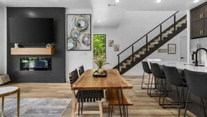 A modern living room with a wooden dining table, black chairs, a flat-screen TV, and a staircase leading to a kitchen area. - Elevated Chalet with Hot Tub Scenic Views & Trail Access (Bella Vista)