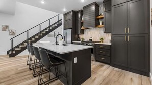 A modern kitchen with dark cabinetry, a central island, and a staircase leading to another level. - Elevated Chalet with Hot Tub Scenic Views & Trail Access (Bella Vista)