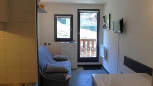 Una habitación compacta con cama, una kitchenette pequeña, un ventanal con vista a un balcón y un televisor montado.