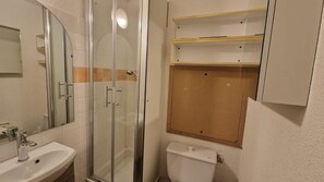 A bathroom with a glass-enclosed shower, a white sink, and a toilet. There are wooden shelves above the toilet.