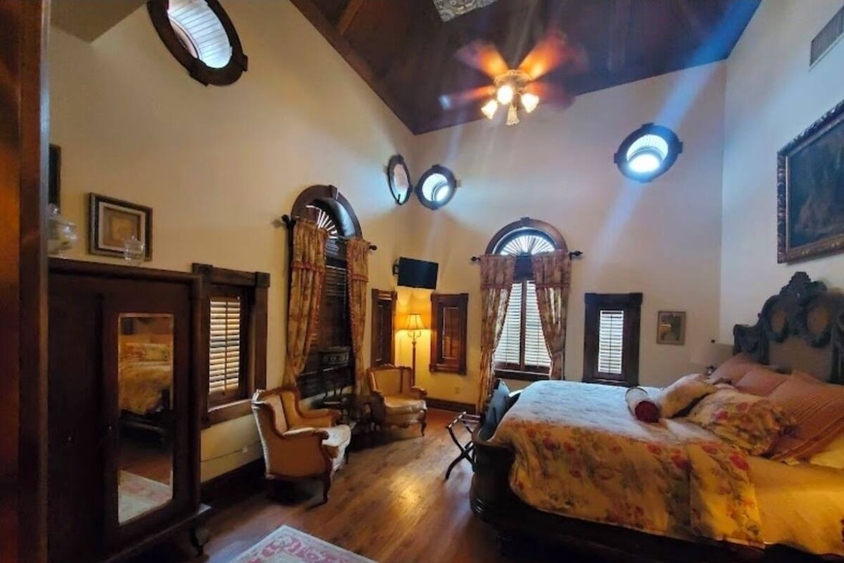 Une chambre à coucher avec un grand lit, un plancher en bois, un ventilateur de plafond et de nombreuses fenêtres avec des rideaux.