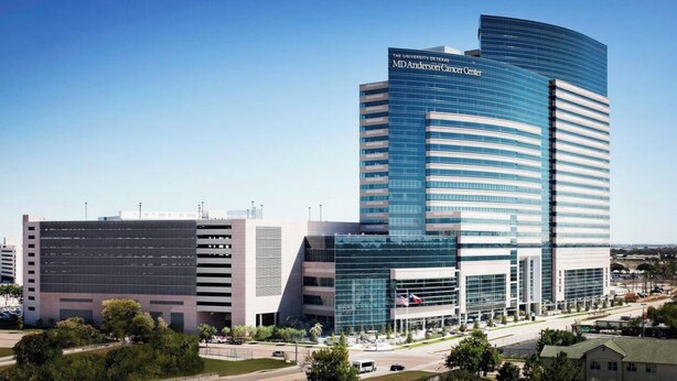 Moderni rakennus, jossa on kaareva lasijulkisivu, sen yläpuolella näkyvä suuri kyltti, jossa lukee ”MD Anderson Cancer Center”, ja sen edessä sijaitseva parkkipaikka.