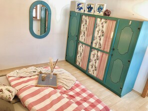 Une chambre à coucher avec un lit, un miroir, une commode avec des rideaux à motifs floraux, et un plateau avec une tasse et une bougie.