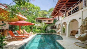 A swimming pool with lounge chairs and umbrellas, surrounded by greenery and a building with arches. - Jungle Villa w/ Lush Gardens walk 2 Pelada Beach (Nosara)