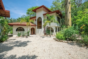 A two-story house with a gravel driveway, surrounded by lush greenery and palm trees.
