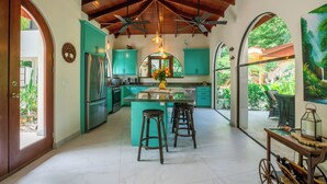 A kitchen with teal cabinets, a central island with stools, ceiling fans, and a view of greenery outside. - Jungle Villa w/ Lush Gardens walk 2 Pelada Beach (Nosara)