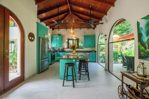 A kitchen with teal cabinets, a central island with stools, ceiling fans, and a view of greenery outside.
