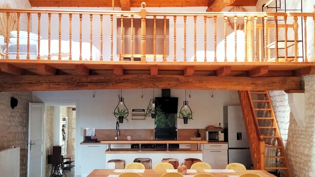 Un loft spacieux avec un plafond en bois, un coin repas avec des chaises jaunes, une cuisine et un coin salon confortable avec un canapé et des coussins.