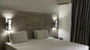 Hotellihuone, jossa on suuri sänky, kaksi seinälle asennettua valaisinta, ilmastointilaite ja ikkuna, jossa on verhot.