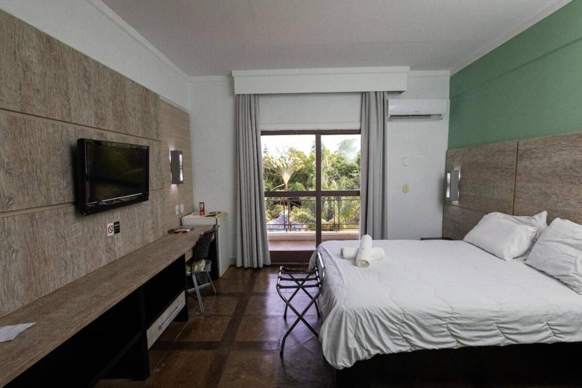 A hotel room with a bed, a TV mounted on a wooden panel wall, a desk with a chair, and a balcony with a view of greenery.