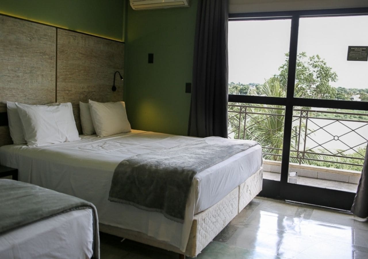 A hotel room with a bed, a bedside table, a window with a view of greenery, and a balcony with a railing.