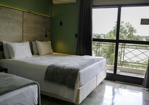 A hotel room with a bed, a bedside table, a window with a view of greenery, and a balcony with a railing. - Araguaia Hotel (Barra do Garcas)